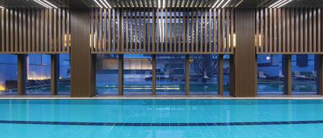 A modern indoor swimming pool with a geometric tile pattern, large windows, and a contemporary wooden ceiling design.