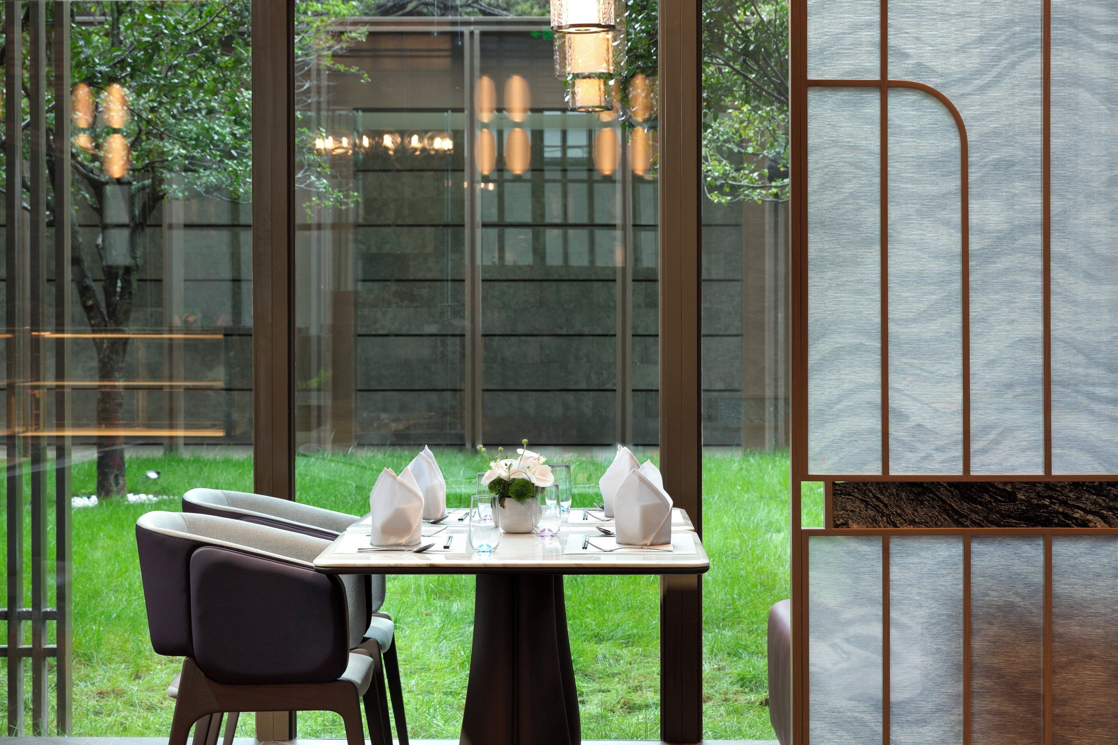 A dining area with a table set for two, a vase with flowers, and a view of a garden through large windows.