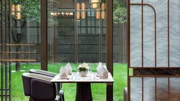 A dining area with a table set for two, a vase with flowers, and a view of a garden through large windows.