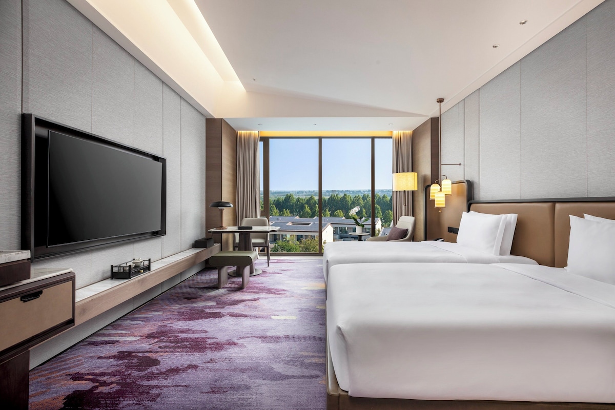 Habitación de hotel moderna con un televisor de pantalla plana grande, dos camas, un escritorio y vistas a la ciudad a través de amplios ventanales.