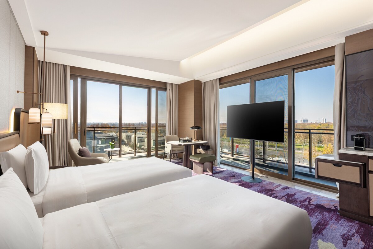 Ein Hotelzimmer mit zwei Betten, einem Schreibtisch, einem Sessel, einem Fernseher und großen Fenstern mit Blick auf die Stadt.