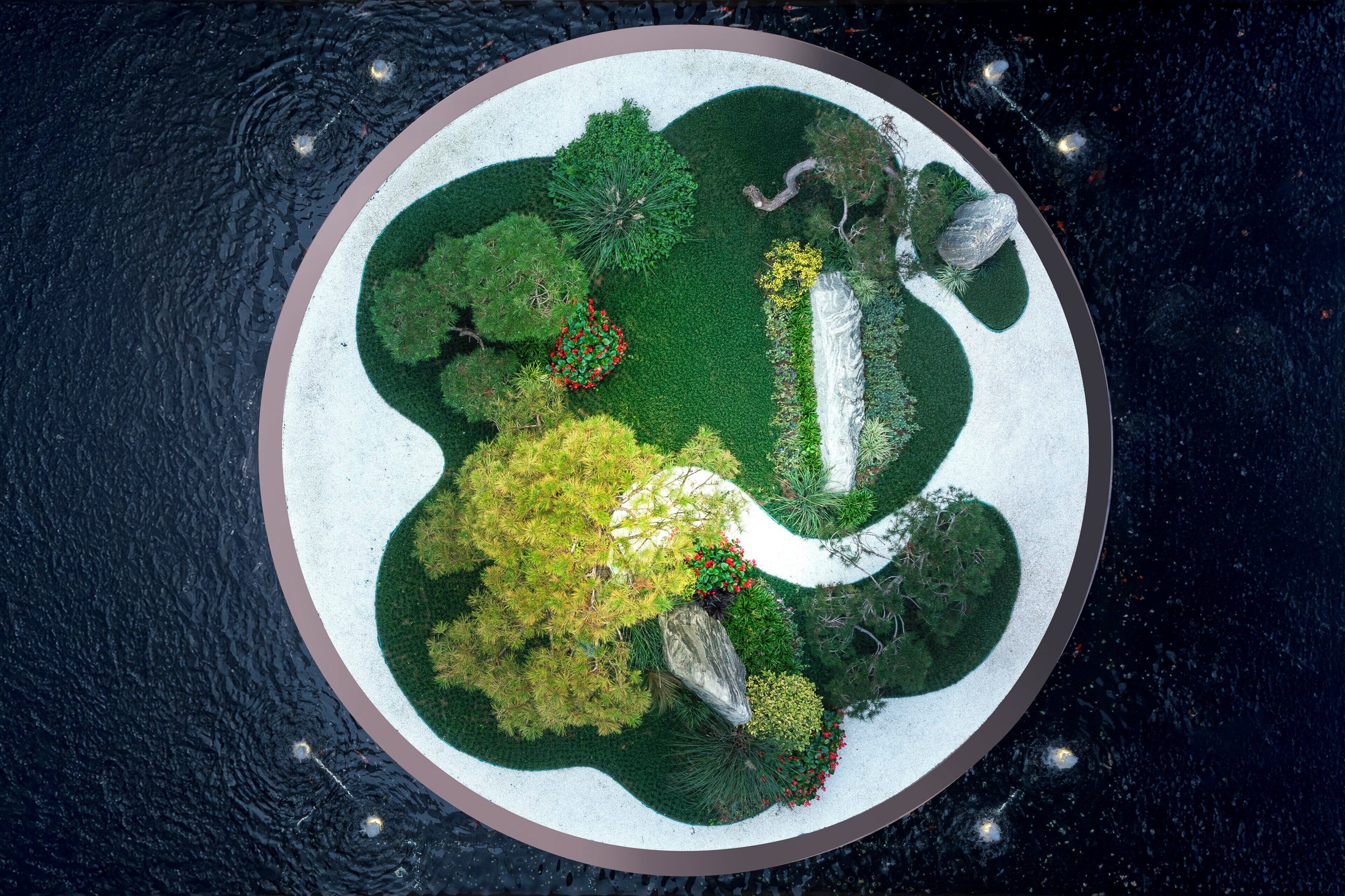 Una vista aerea di un giardino circolare con diversi alberi e arbusti, circondato da uno specchio d'acqua.