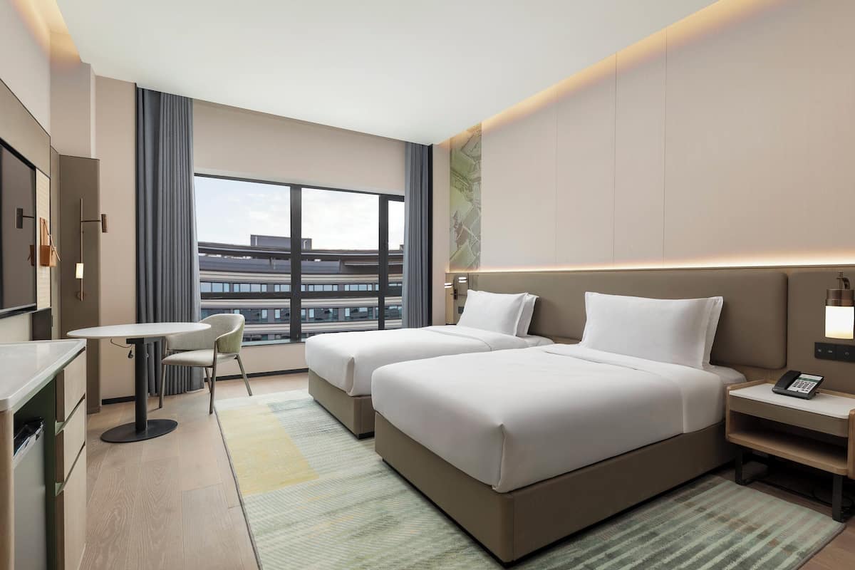A hotel room with two beds, a small table, a chair, a desk, and a view of the cityscape.