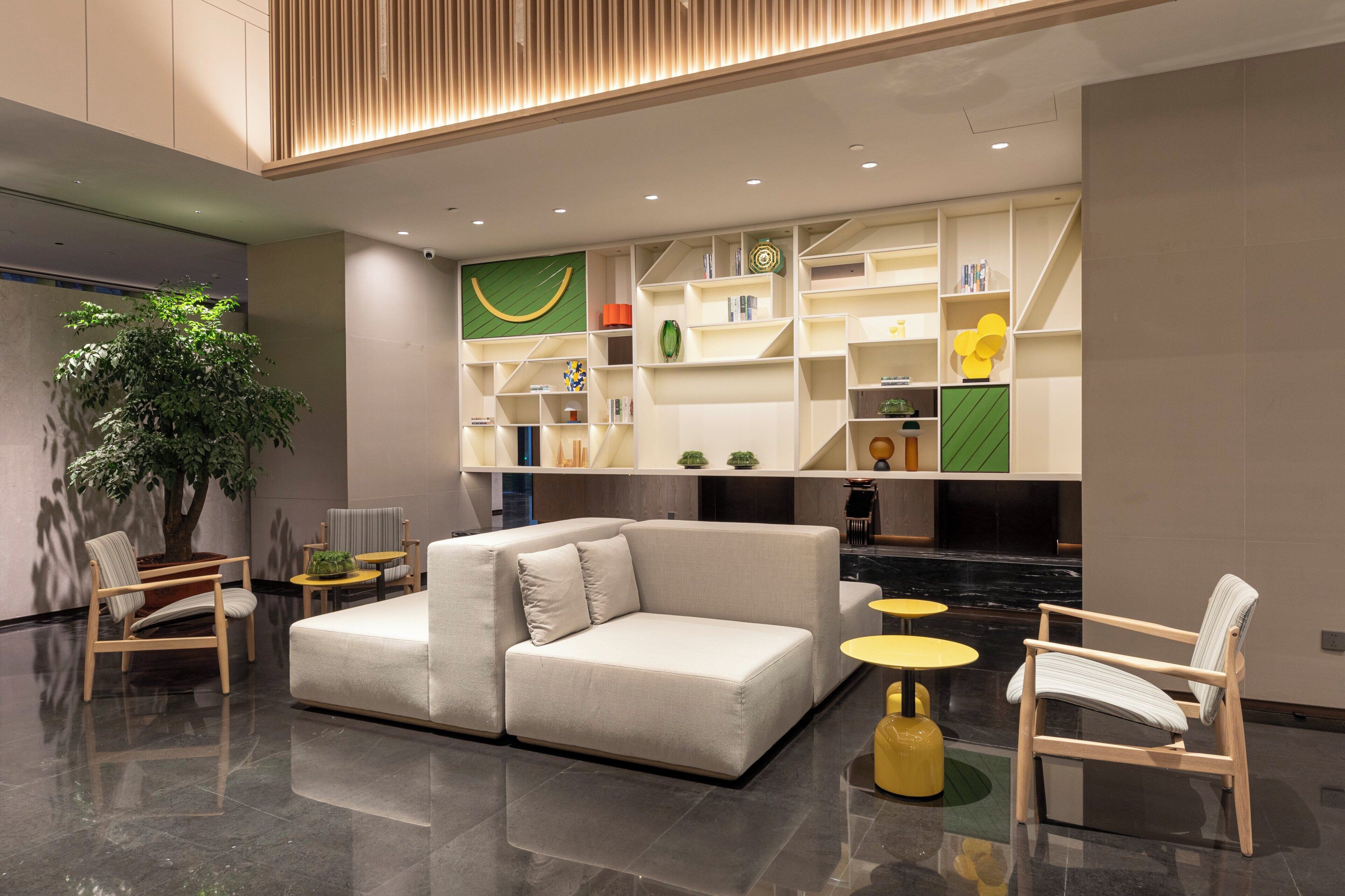 A modern lobby with a sectional sofa, wooden chairs, a potted plant, and a wall with built-in shelves containing various items.