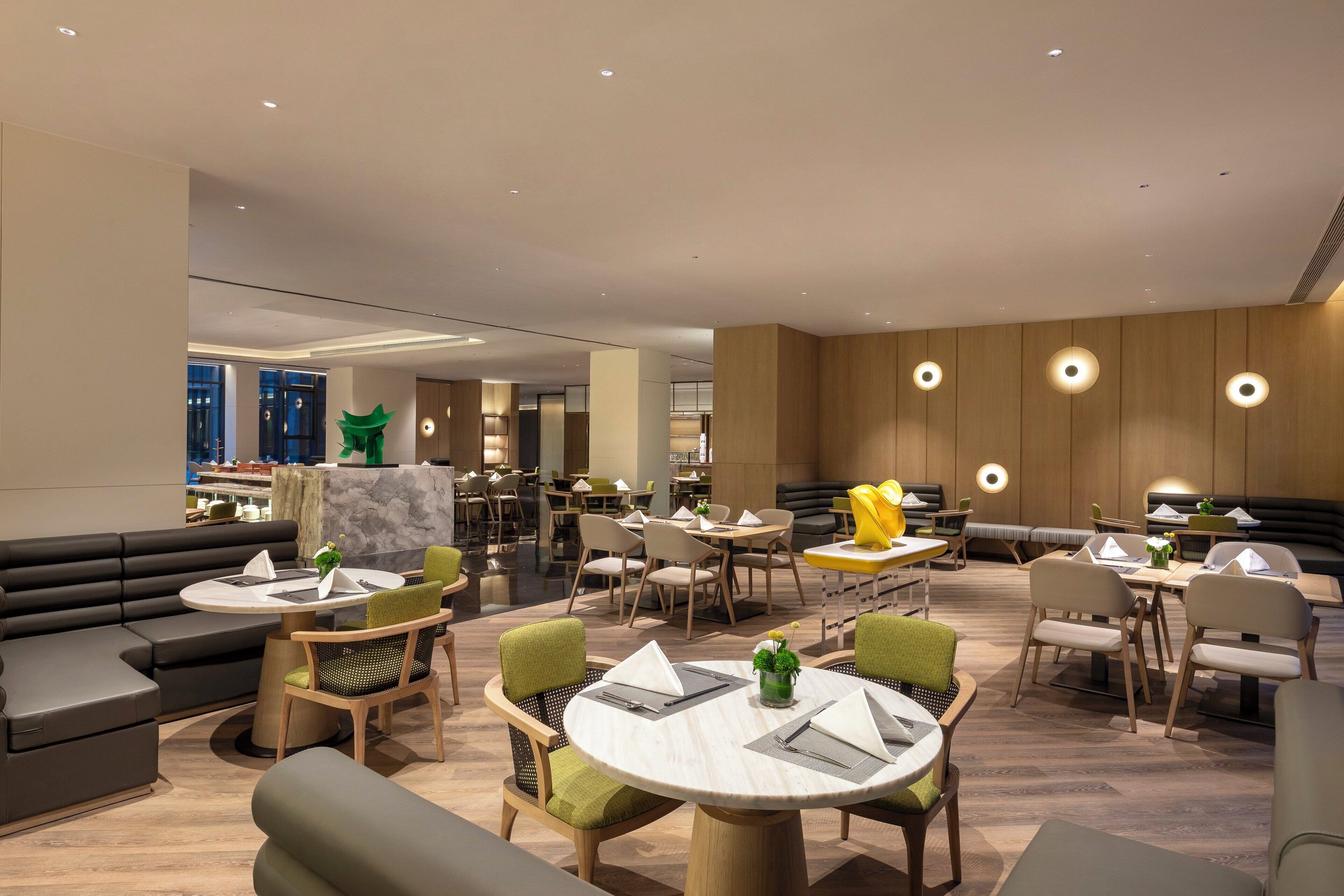 Un hall d’hôtel moderne doté de plusieurs espaces de détente, notamment des canapés et des tables rondes, et un bar bien visible au fond.