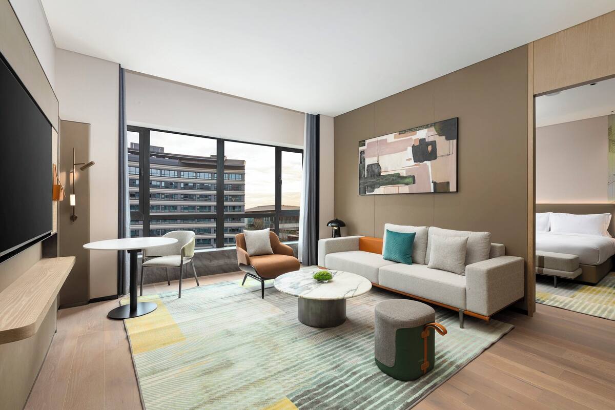 A modern living room with a sofa, armchair, coffee table, and a large window offering a city view.