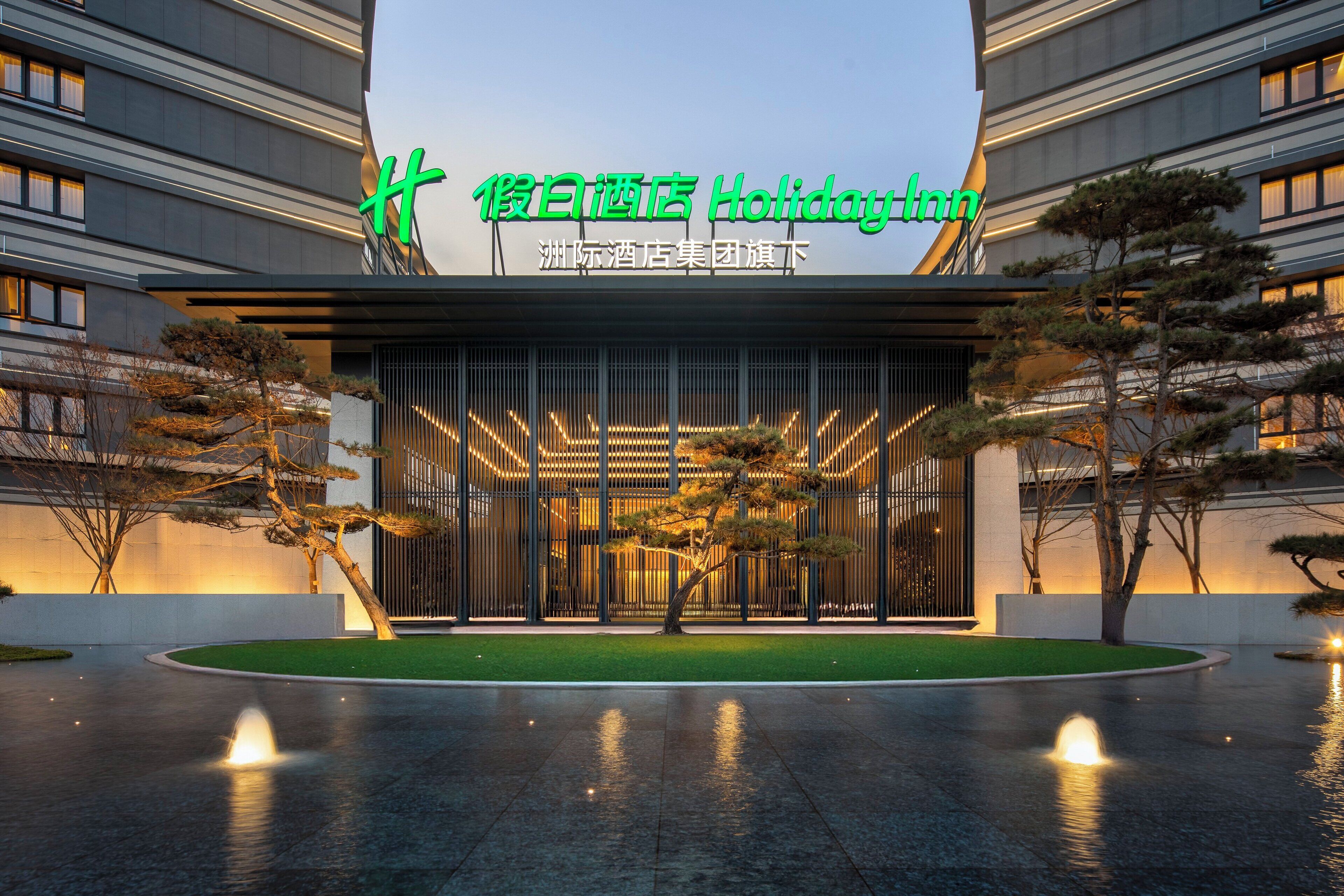 A modern hotel entrance with a large illuminated sign, a well-maintained lawn, and a water feature in front.