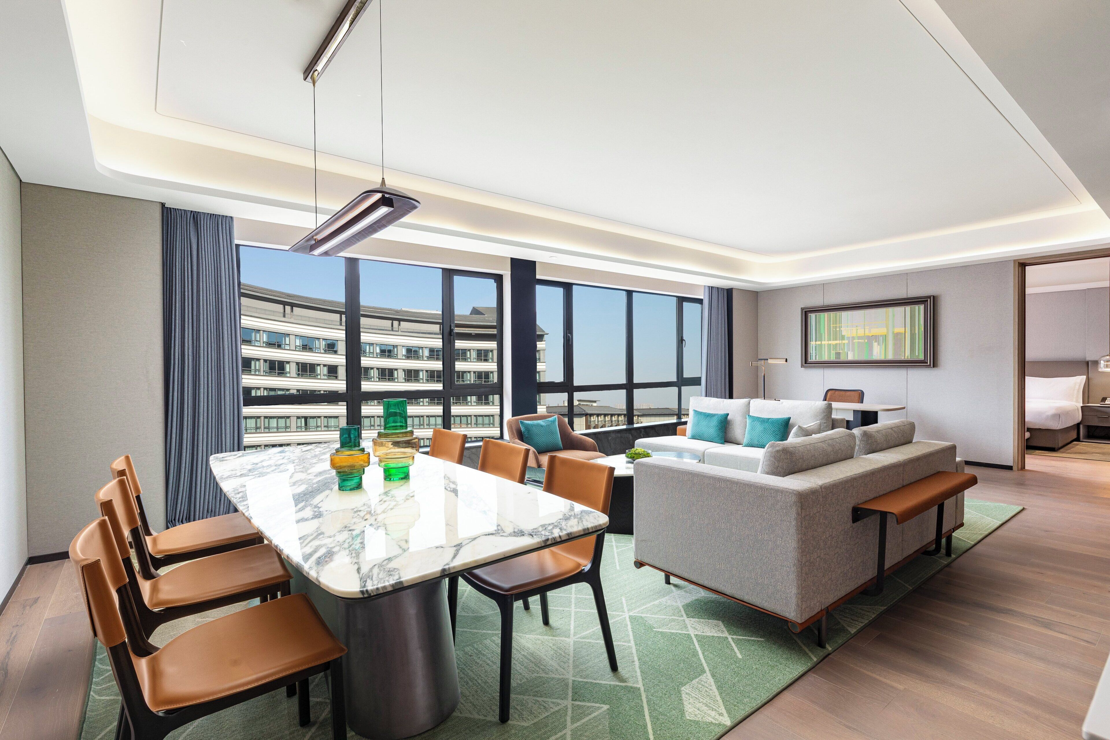 A modern hotel room with a dining area, a sofa, and a large window offering a city view.