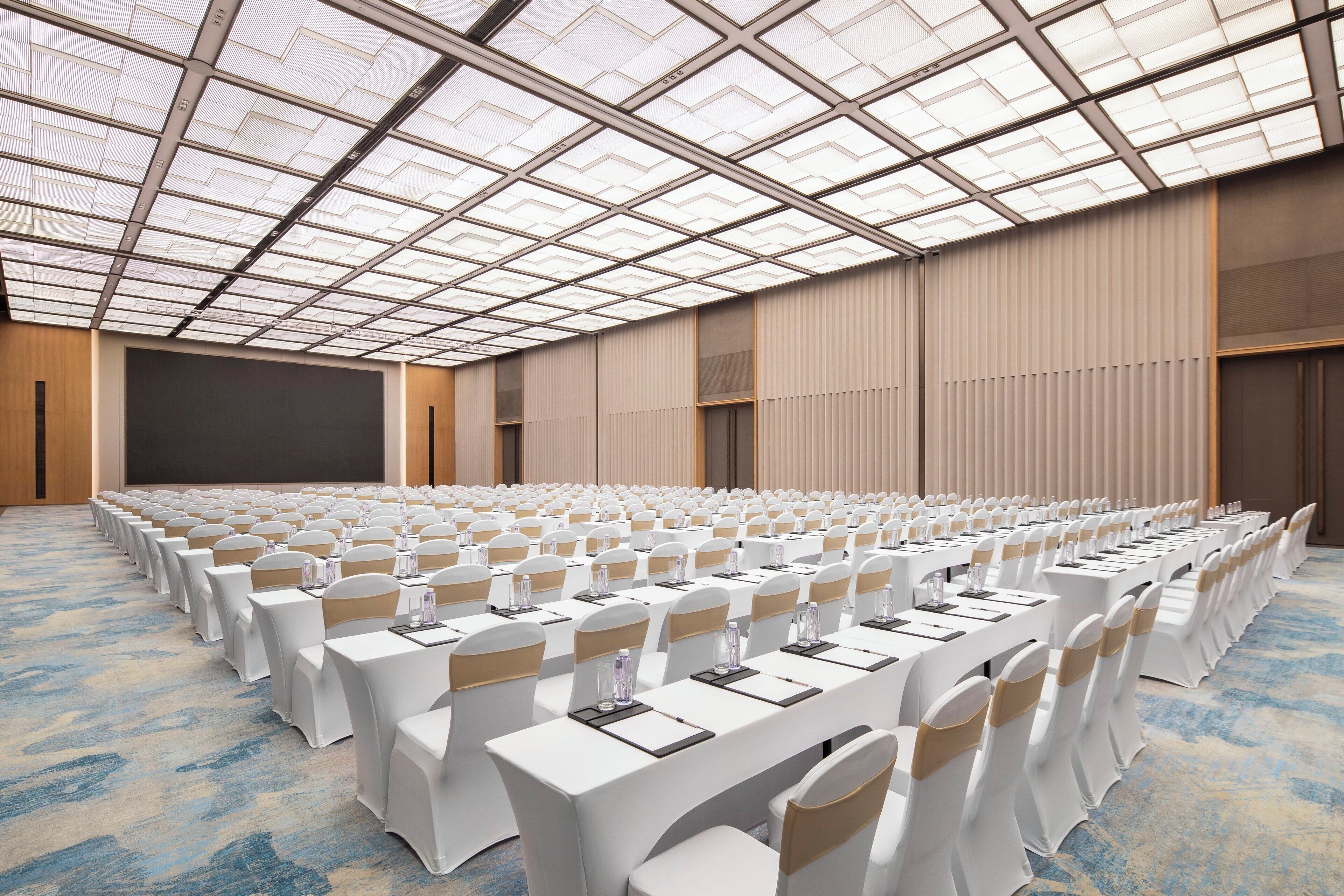Une salle de conférence avec des rangées de tables et de chaises blanches, un grand écran et un plafond haut doté de lucarnes.