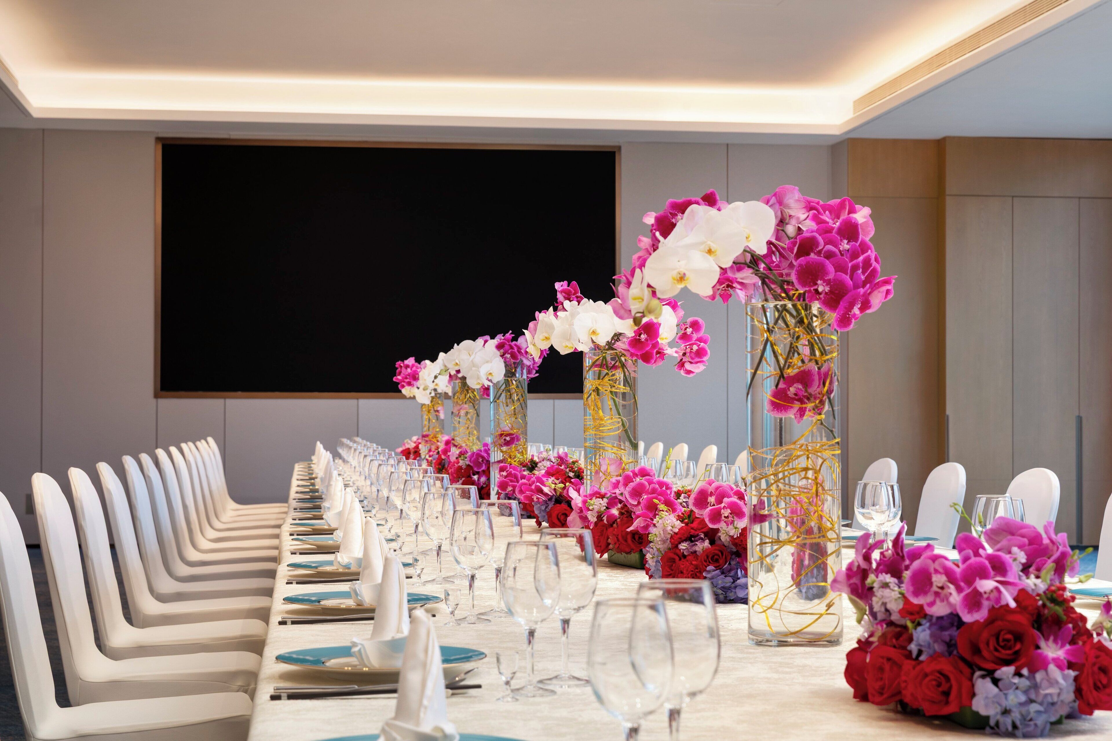 Mesa longa posta para um evento formal, com cadeiras e taças de vinho, decorada com arranjos florais.