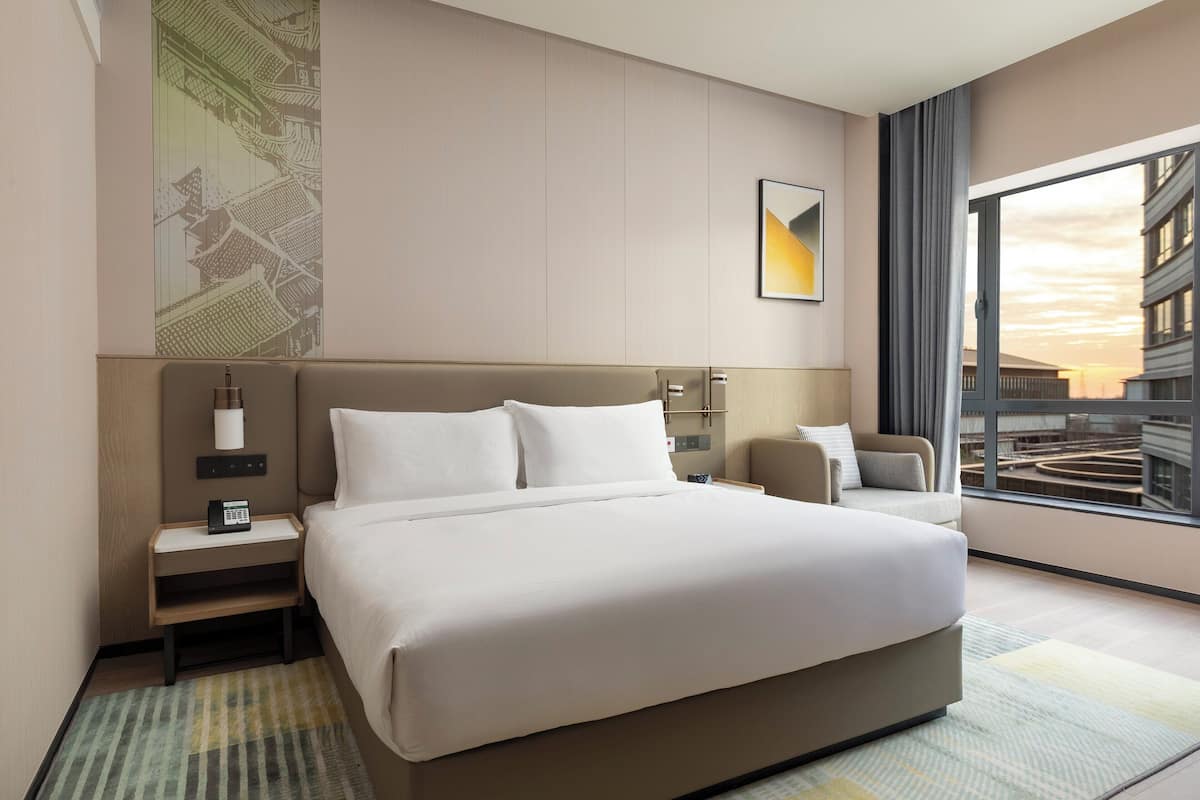 A hotel room with a large bed, a nightstand, a lamp, a chair, and a large window with a view of buildings.