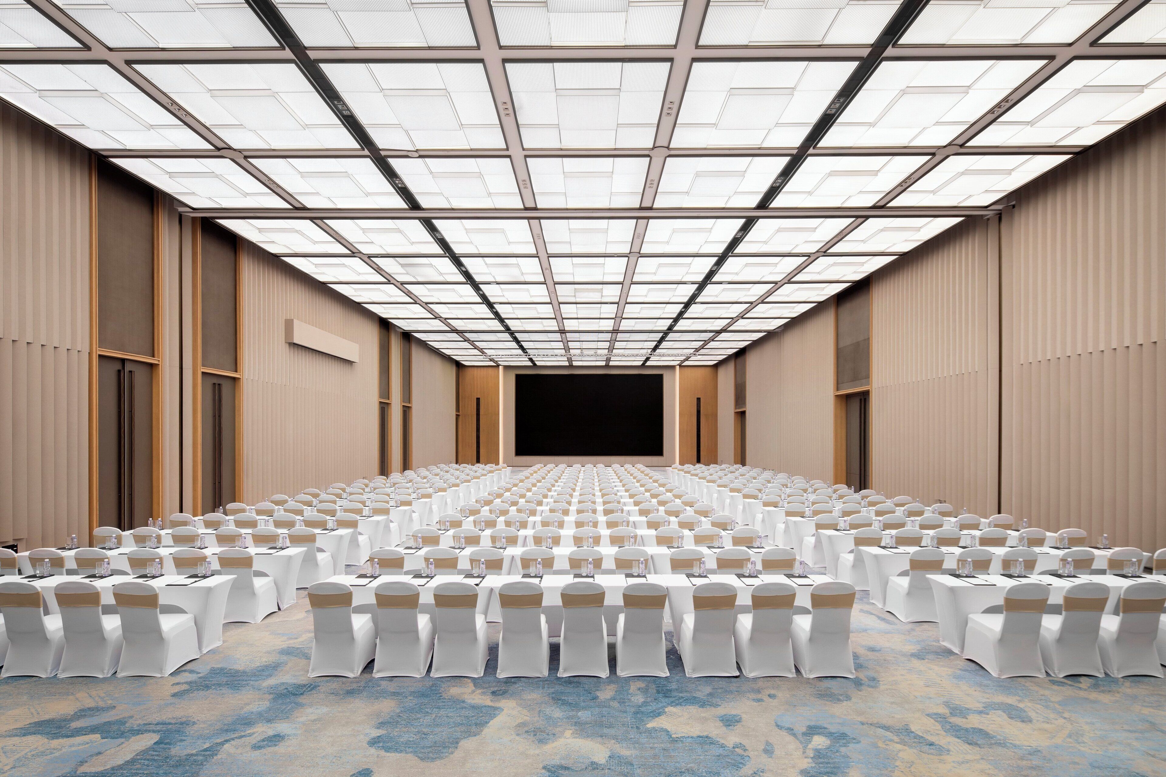 A large conference room with rows of tables and chairs, a stage at the front, and a high ceiling with skylights.