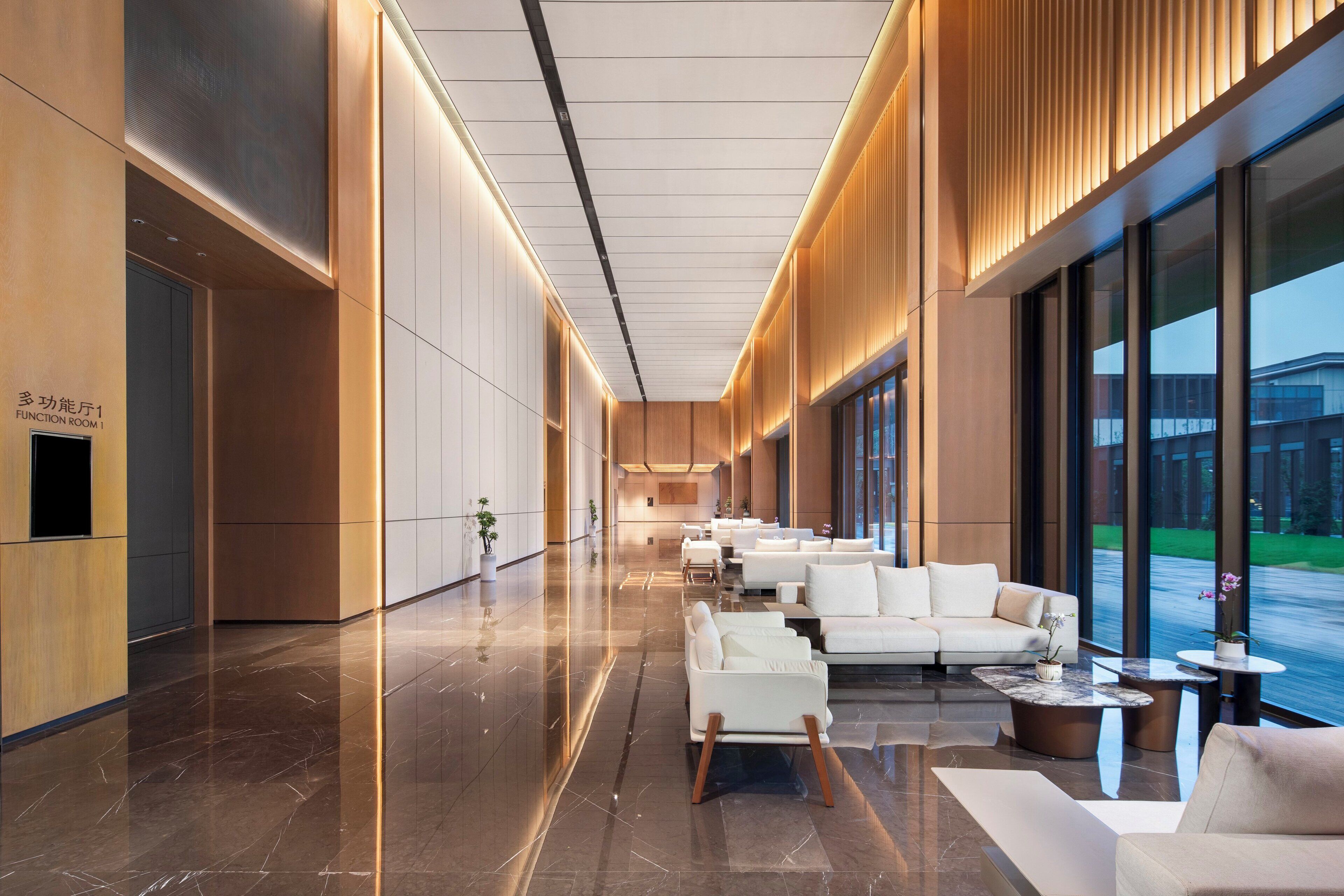 A modern hotel lobby with a long corridor, seating area with sofas and tables, and large windows offering views of the outside.