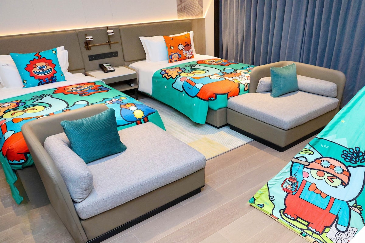 A hotel room with two beds, each with a colorful cartoon-themed bedspread, two gray sofas with blue cushions, a small table with a phone, and a tent-like structure with a cartoon character.