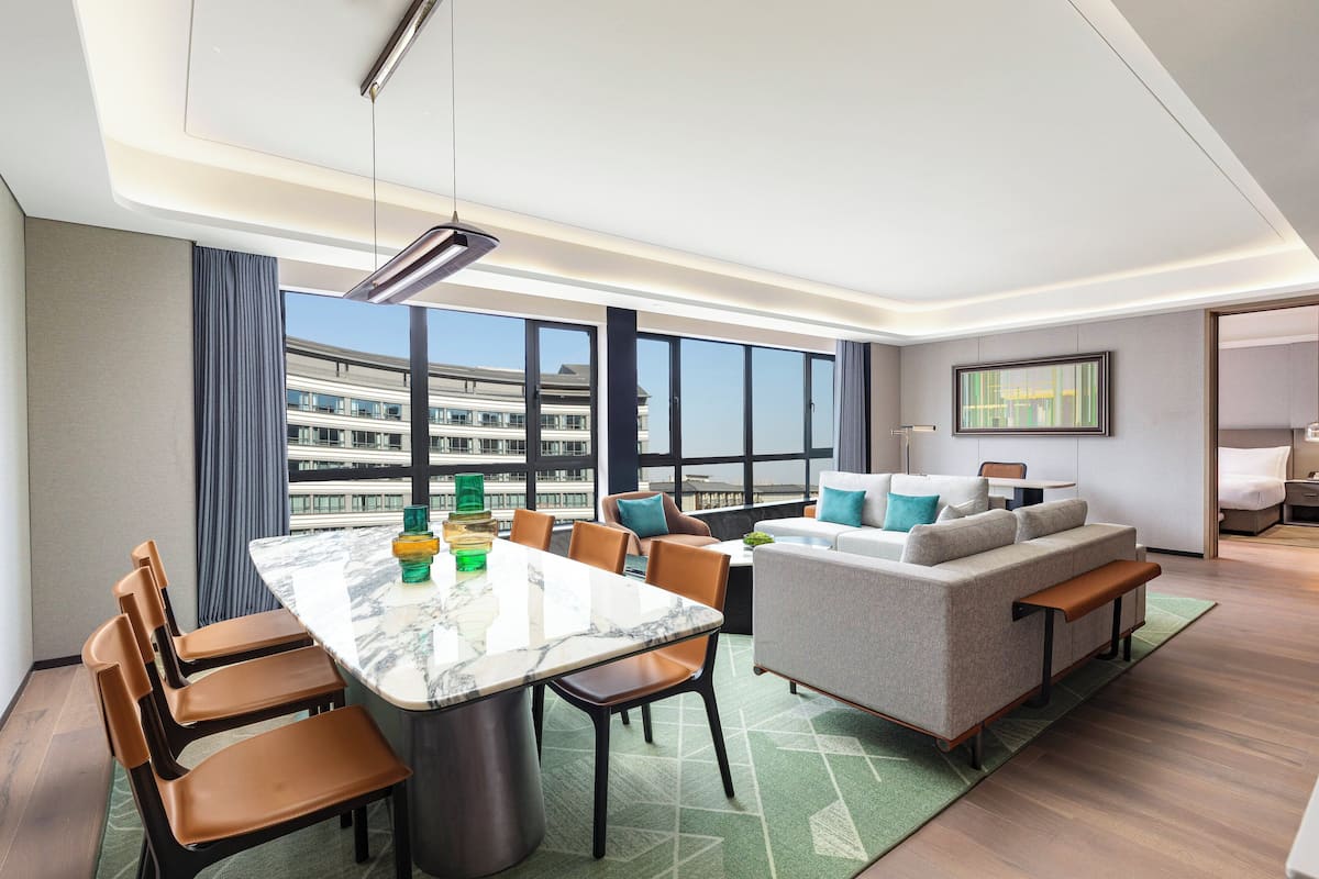 A modern hotel room with a dining area, a sofa, and a large window offering a city view.