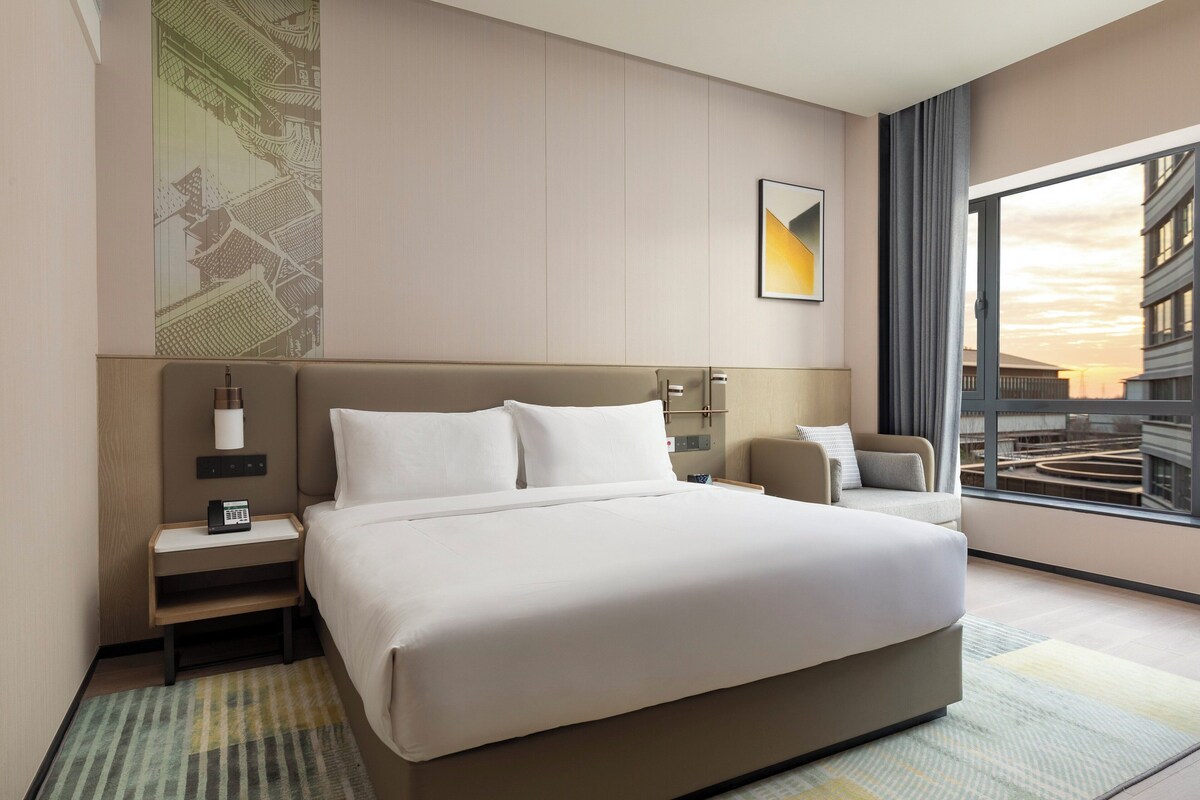 Una camera d'albergo con un letto grande, un comodino, una lampada, una sedia e un'ampia finestra con vista sugli edifici.