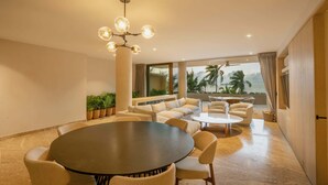 Uma área de jantar moderna com uma mesa redonda e cadeiras, um conjunto de sofás e uma janela ampla com vista para palmeiras e um corpo d'água.