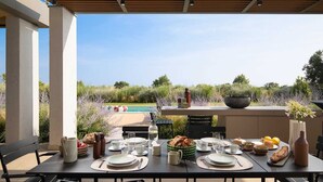 Ett utomhusområde med plats för att äta, med utsikt över en swimmingpool, omgivet av grönska och en träpergola.