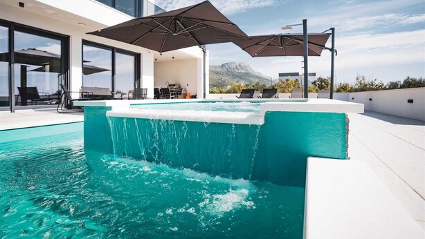 Un espace piscine extérieur moderne, agrémenté d’une cascade, entouré de chaises longues et de parasols, le tout se fondant dans un paysage montagneux.