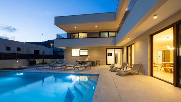 A modern house with a swimming pool, outdoor seating, and a well-lit interior. - Villa Veluno with Pool (Kaštel Lukšić)
