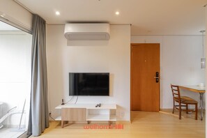 Ein modernes Wohnzimmer mit einem Flachbildfernseher, der an einem weißen Tischchen befestigt ist, einer Decken-Klimaanlage und einer Holztür im Hintergrund.