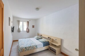 A bedroom with a bed, a bedside table, a window with curtains, and a wall-mounted light fixture. - A peaceful getaway in Corsica—private garden, 5 minutes from the beach. (Aléria)