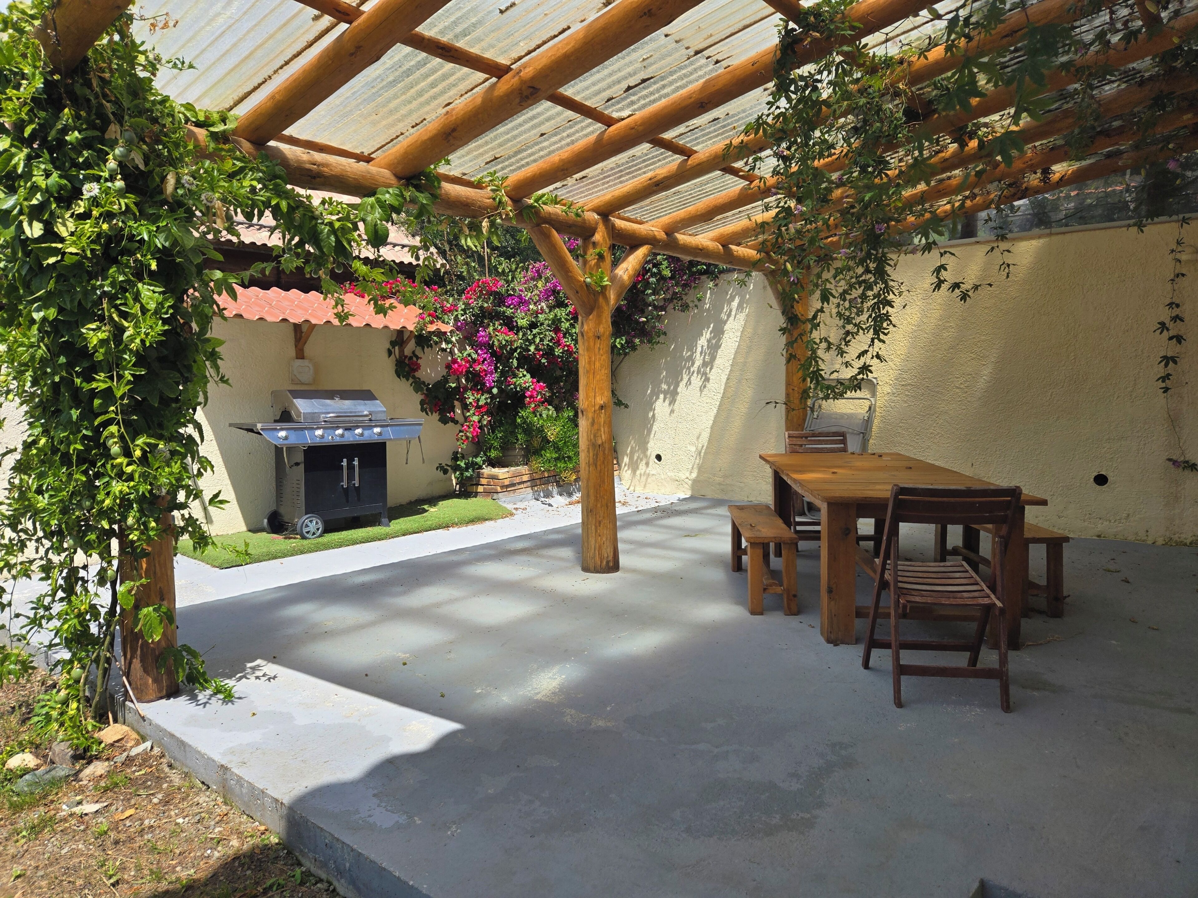 Eine Terrasse im Freien mit einem Holztisch, Bänken, einem Grill und einer Pergola.