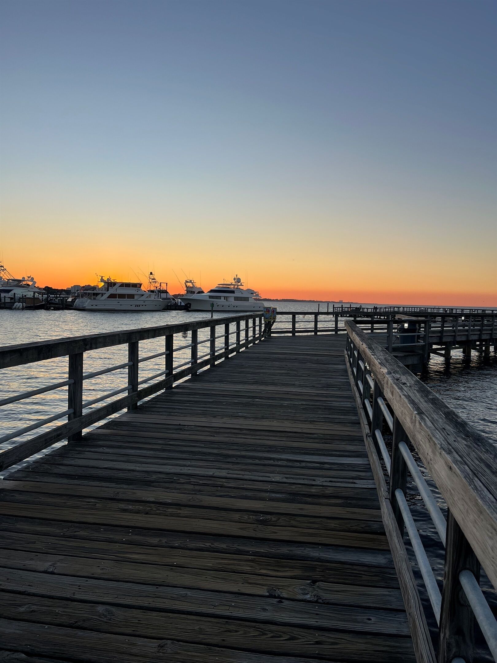 A wooden pier extending into calm waters at sunset, with several yachts docked nearby.