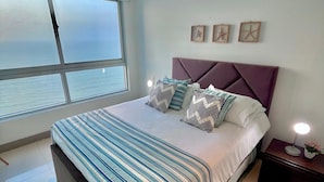 Ein modernes Schlafzimmer mit einem großen Bett, Meerblick durch ein Fenster und einem Nachttisch mit Lampe.