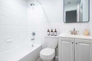 Ett modernt badrum med vita kakelplattor, ett badkar, en toalett och ett handfat med tillhörande hygienartiklar.