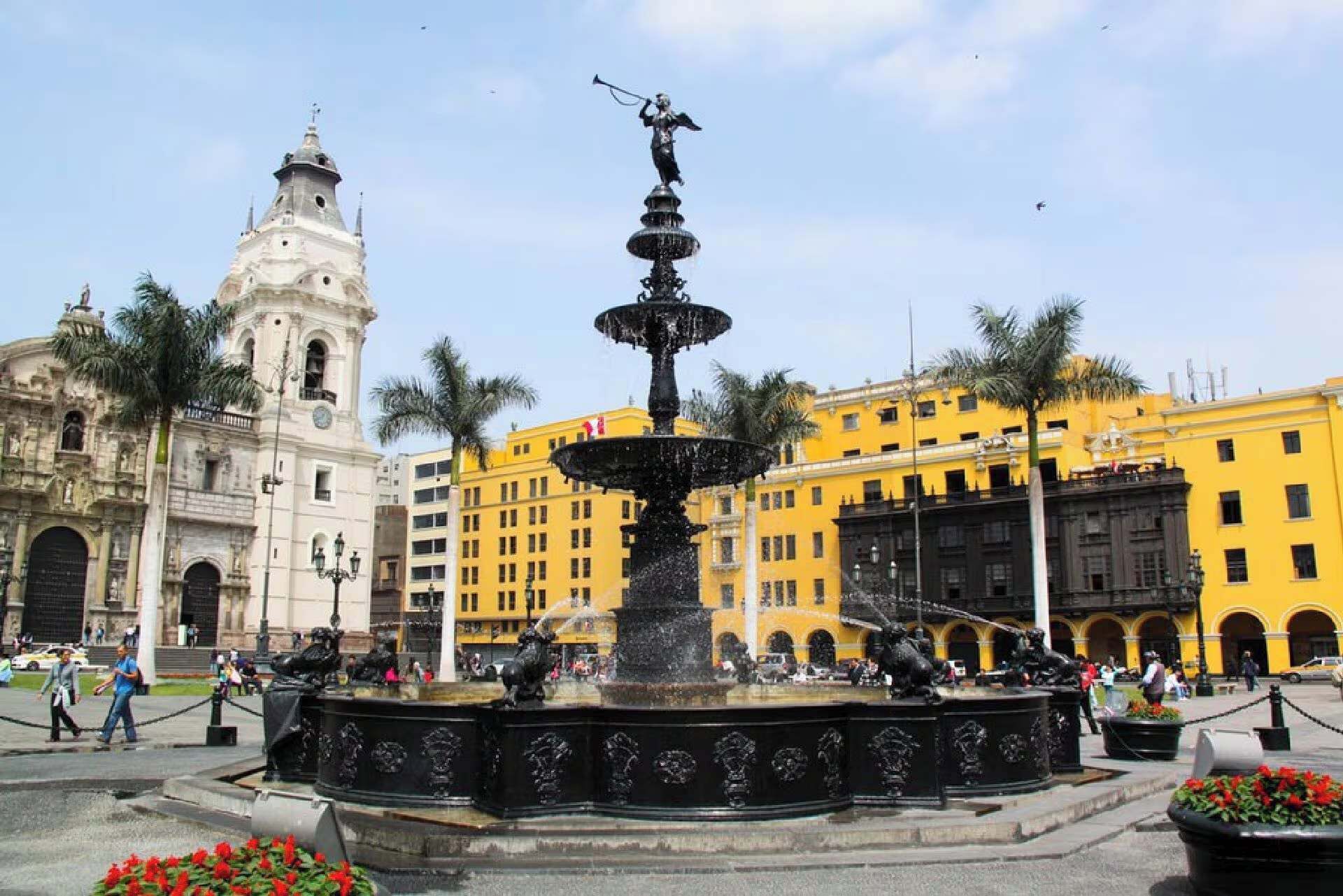 Un bâtiment historique doté d’une tour horlogée, d’une fontaine ornée de statues et d’une place bordée d’immeubles jaunes.