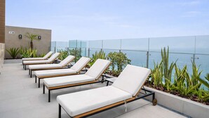 A rooftop lounge area with white cushioned lounge chairs arranged in a row, overlooking a body of water. - Sight 1707 · Ocean View 2BR Stylish Apt • Pool, Gym & Parking (Lima)