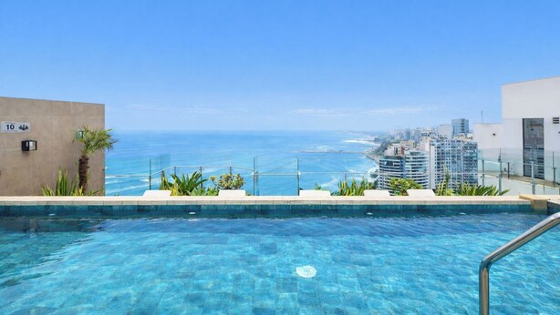 A pool with clear blue water, surrounded by plants and a view of the ocean and cityscape. - Sight 1707 · Ocean View 2BR Stylish Apt • Pool, Gym & Parking (Lima)