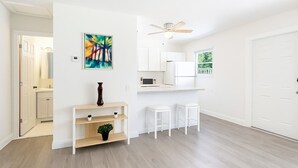 Une cuisine moderne avec des armoires blanches, un four à micro-ondes et un ventilateur de plafond. On y trouve un petit coin salon avec des tabourets et une étagère ornée d'une plante.