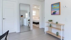 Un couloir avec une porte blanche, un miroir qui reflète une pièce où se trouve un lit, et une étagère ornée d’objets décoratifs.