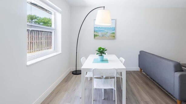 Een kamer met een witte tafel, twee witte stoelen, een grijze bank, een vloerlamp en een schilderij aan de muur.