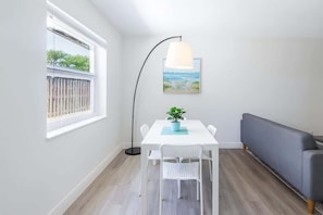 Una habitación con una mesa blanca, dos sillas blancas, un sofá gris, una lámpara de pie y un cuadro en la pared.