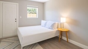 Een slaapkamer met een enkel bed, een nachtkastje met een lamp, een raam en een deur.