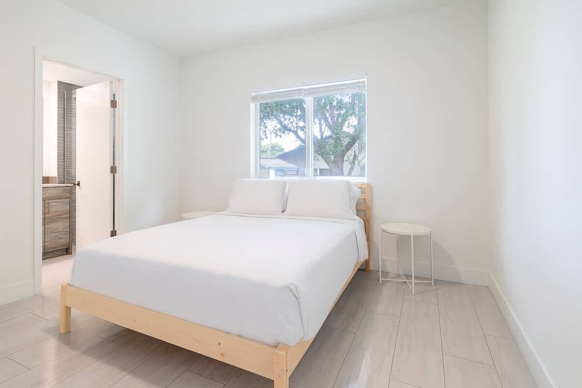 Ein schlicht eingerichtetes Schlafzimmer mit einem hölzernen Bett, einem kleinen Nachttisch und einem Fenster mit Blick auf Bäume.