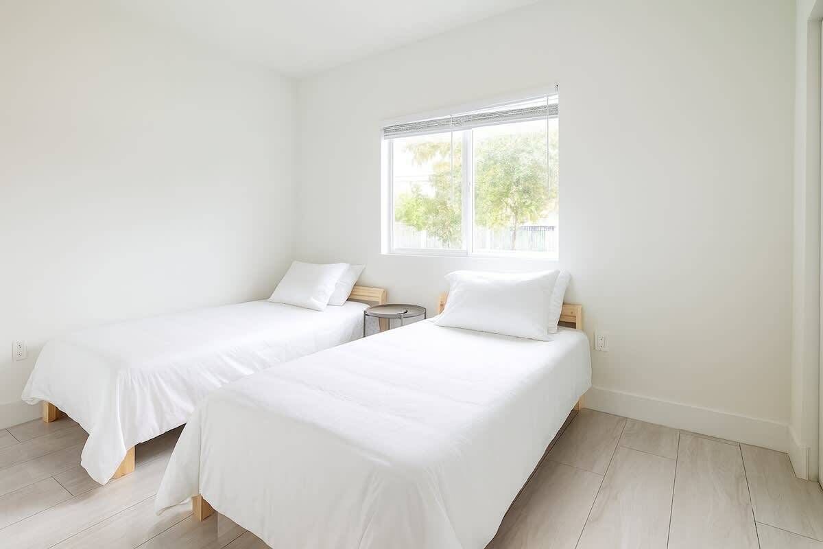 Een kamer met twee eenpersoonsbedden, een klein nachtkastje, een raam met zonwering en een licht houten vloer.