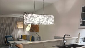 Eine moderne Küche mit weißer Arbeitsplatte, integriertem Geschirrspüler und Spüle aus Edelstahl. Zwei grüne Barhocker und eine Pendelleuchte mit geometrischem Design sind ebenfalls vorhanden.