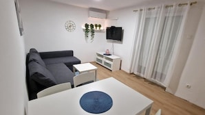A small, modern living room with a gray sofa, a white table with a blue placemat, a TV on a white stand, and a window with sheer curtains. - Apartments Rivijera 1 (Crikvenica)