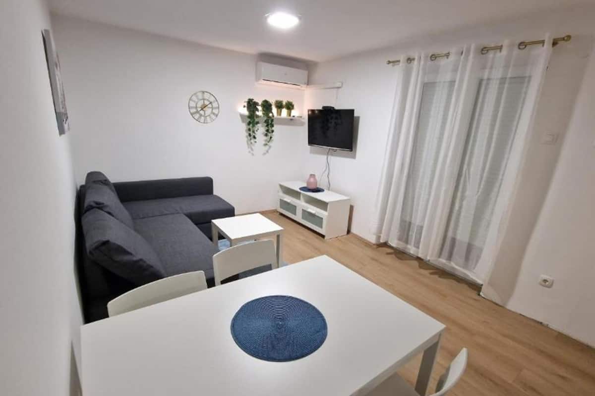 A small, modern living room with a gray sofa, a white table with a blue placemat, a TV on a white stand, and a window with sheer curtains.