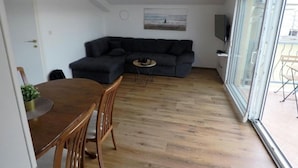 A living room with a dark-colored sofa, a small round table, and a dining table with chairs. There is a large window with a view of the outside. - Apartments Rivijera 1 (Crikvenica)