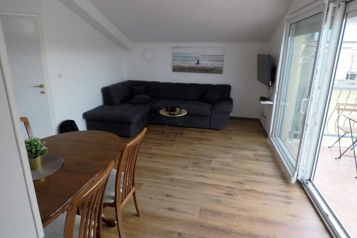 A living room with a dark-colored sofa, a small round table, and a dining table with chairs. There is a large window with a view of the outside.
