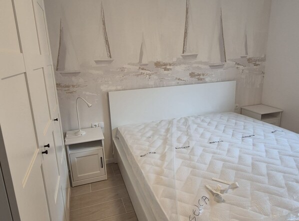 Une chambre à coucher avec un lit blanc, une table de chevet, une lampe et un mur orné d’une fresque représentant un voilier.