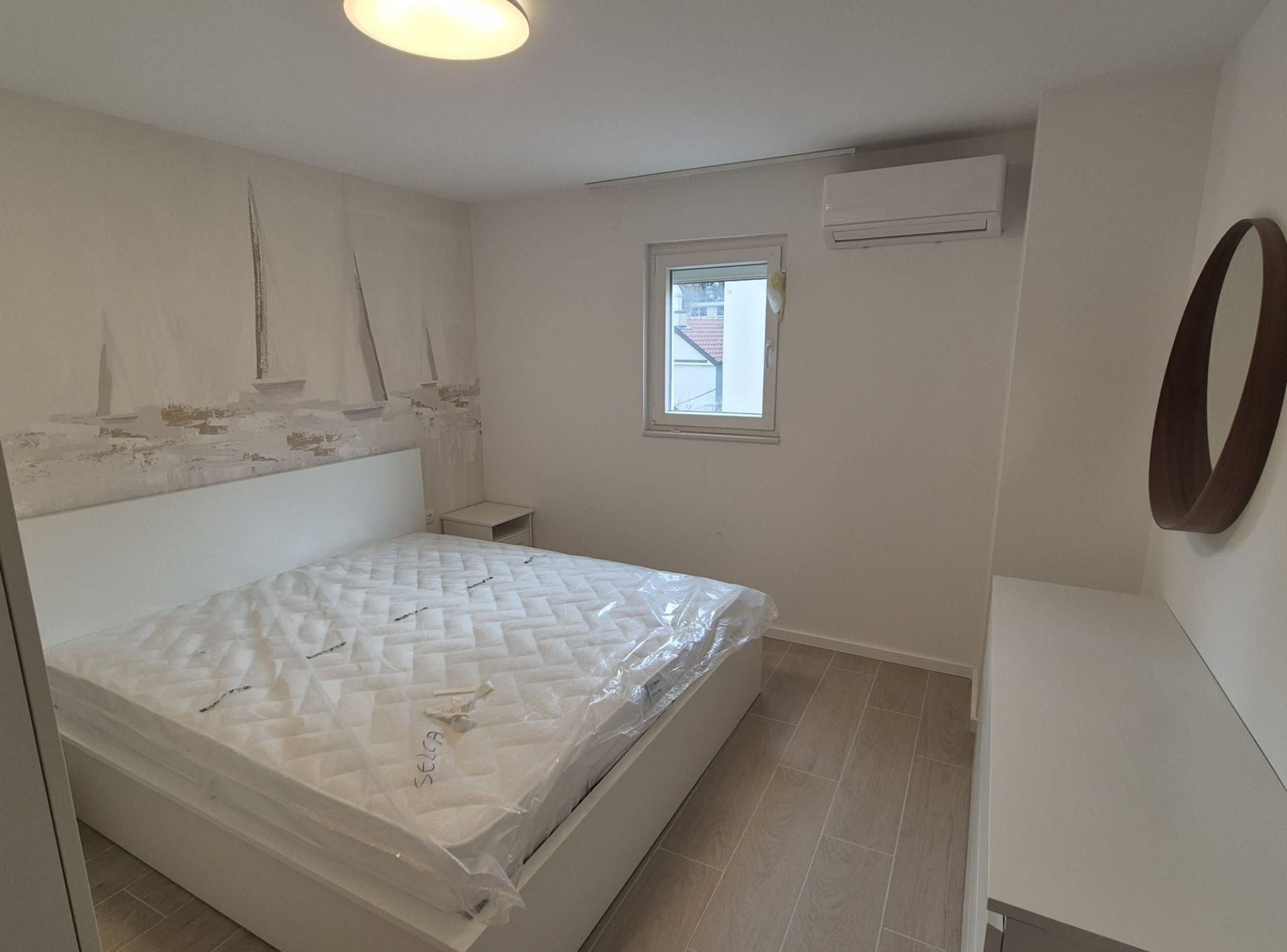 A bedroom with a bed, a small bedside table, a window, and an air conditioning unit.