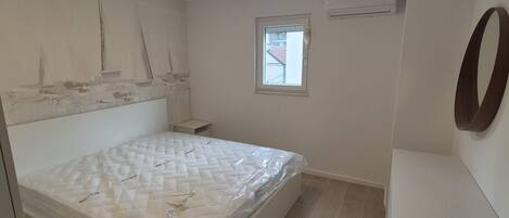 A bedroom with a bed, a small bedside table, a window, and an air conditioning unit.