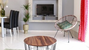 Una sala moderna con una mesa redonda de madera, un sofá negro, un sillón de mimbre con cojín, un televisor sobre una estantería y una planta en maceta.