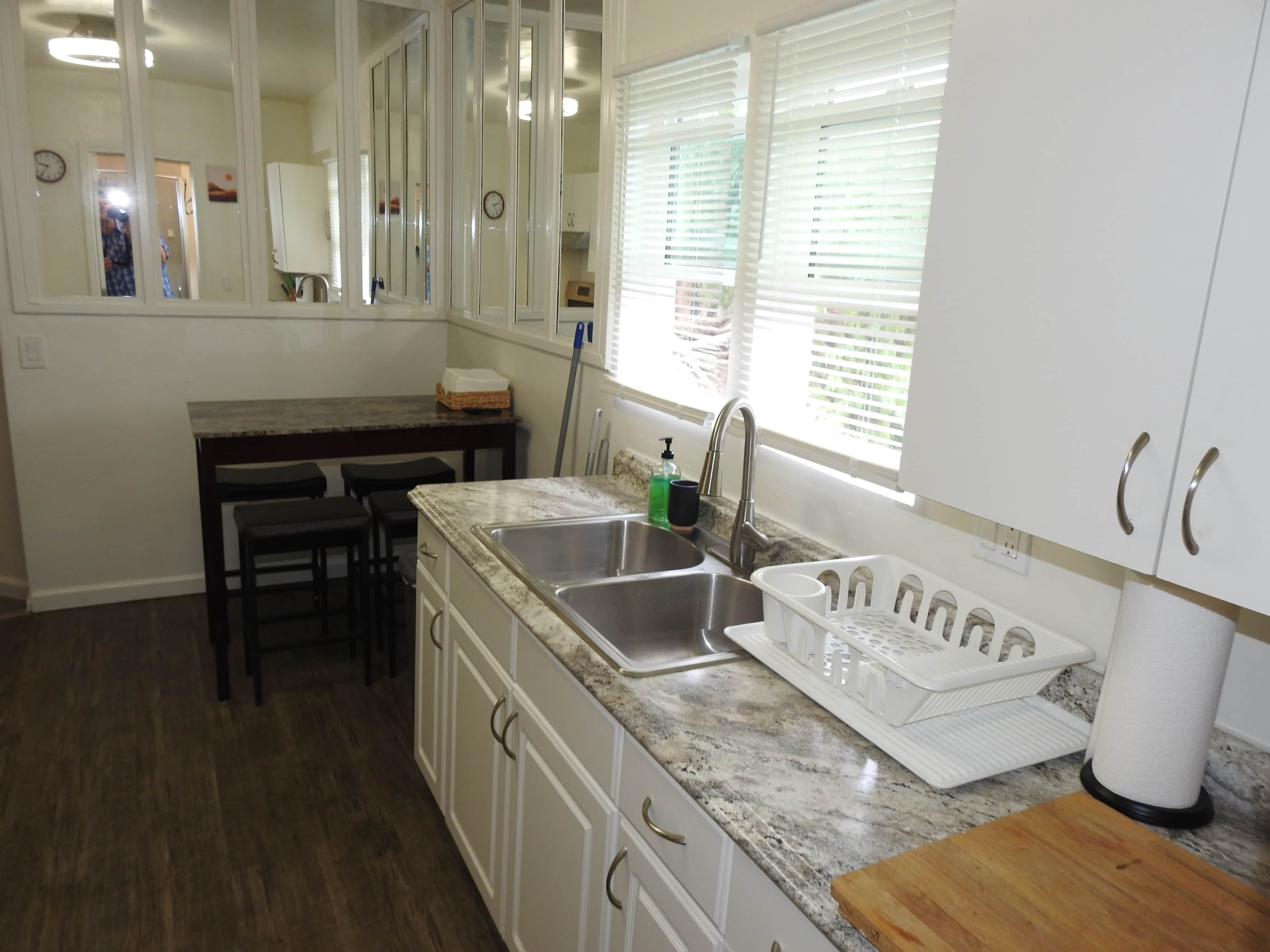 A kitchen with white cabinets, a granite countertop, a sink, a window with blinds, and a wooden cutting board.